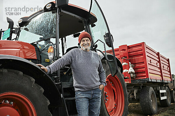 Lächelnder reifer Landwirt steht mit der Hand in der Tasche vor einem roten Traktor auf einem Bauernhof