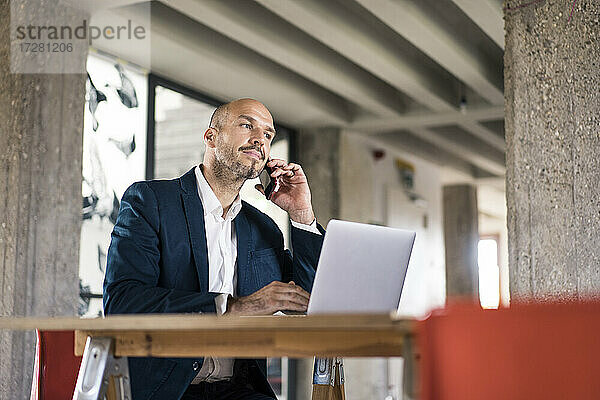 Geschäftsmann im Anzug  der mit seinem Handy telefoniert  während er im Büro am Laptop arbeitet