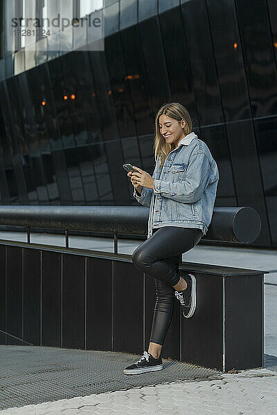 Lächelnde junge Frau  die ihr Smartphone benutzt  während sie sich an ein Geländer vor einem modernen Gebäude lehnt