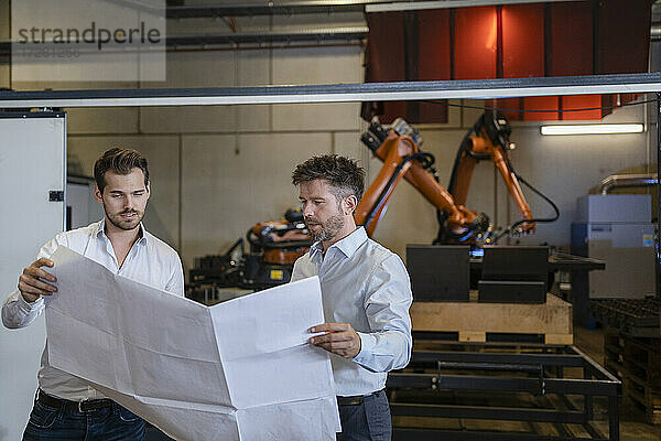 Ein Team mit einer Blaupause diskutiert  während es in einer Fabrik vor einem Roboterarm steht.