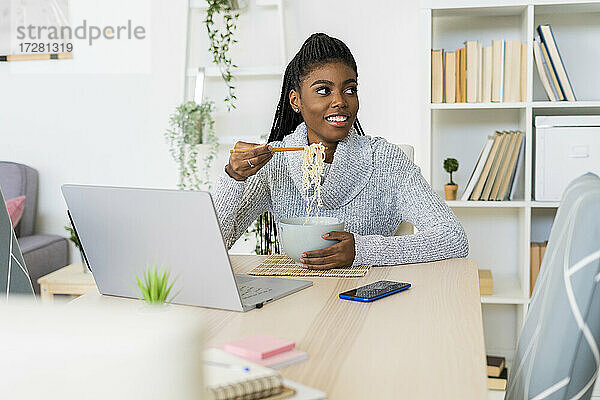 Lächelnde junge Frau  die wegschaut  während sie Nudeln isst und zu Hause sitzt
