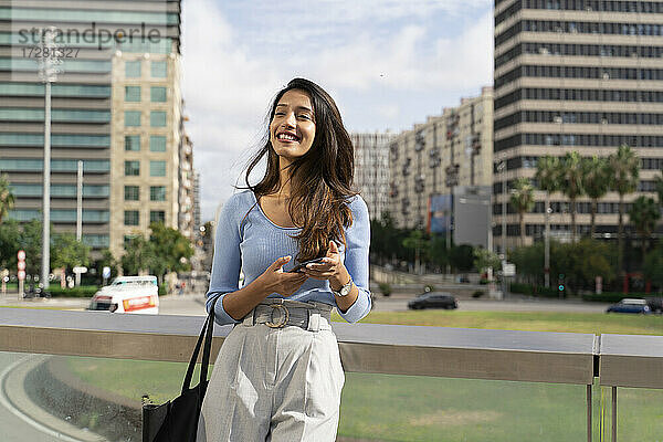 Lächelnde Geschäftsfrau  die ihr Smartphone in der Hand hält und sich an ein Geländer lehnt  mit der Stadt im Hintergrund