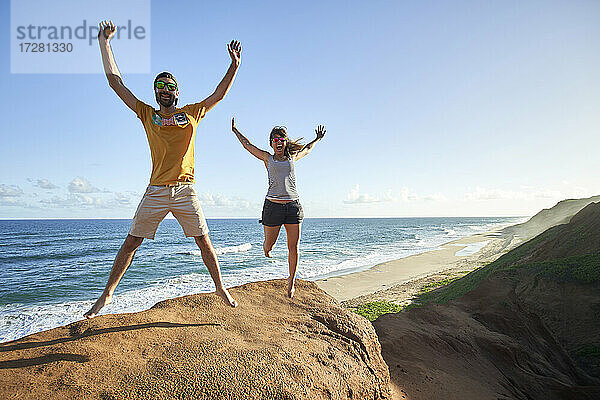 Fröhliches Paar  das mit erhobenen Armen auf eine Felsformation am Strand springt