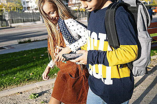 Geschwister halten sich an den Händen  während sie ihr Smartphone benutzen und in einem öffentlichen Park an einem sonnigen Tag spazieren gehen
