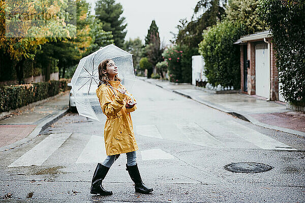 Lächelndes Mädchen im Regenmantel  das einen Regenschirm hält  während es auf einer Straße in der Stadt spazieren geht