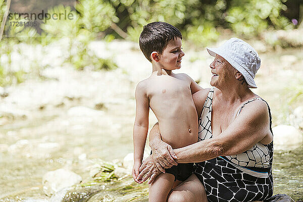 Enkel und Großmutter sitzen lächelnd im Fluss im Wald