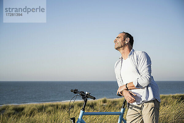 Mann mit geschlossenen Augen lehnt sich am Strand gegen den klaren Himmel auf ein Fahrrad