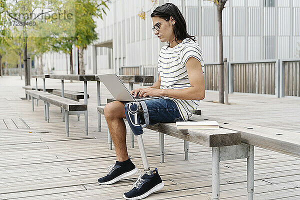 Nachdenklicher Mann mit Beinprothese  der auf einer Bank in der Stadt sitzt und einen Laptop benutzt