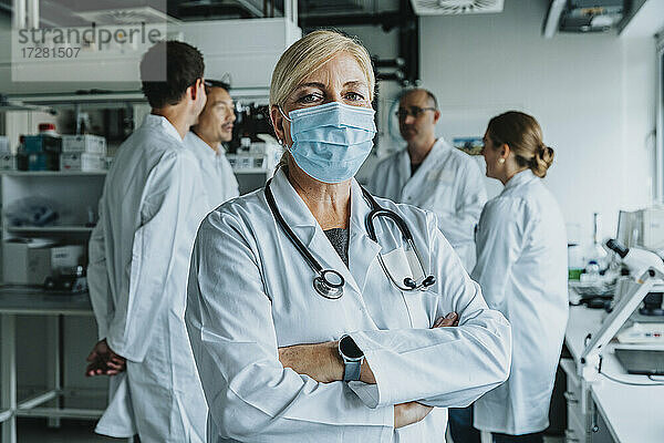 Selbstbewusster Wissenschaftler mit Gesichtsmaske  der mit verschränkten Armen und einem Mitarbeiter im Hintergrund im Labor steht