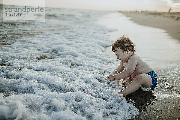Süßer kleiner Junge lachend im Wasser sitzend am Strand bei Sonnenuntergang