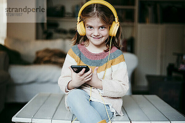 Lächelndes Mädchen mit Kopfhörern  das ein Mobiltelefon benutzt  während es zu Hause auf dem Tisch sitzt