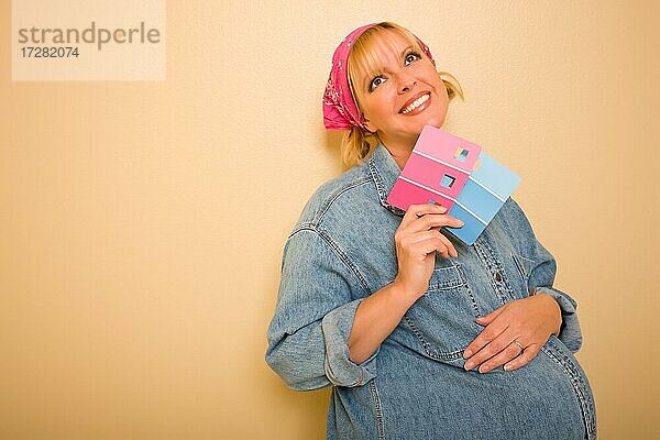 Lächelnd nachdenklich schwangere Frau lehnt an der Wand hält rosa und blauen Farbe swatches