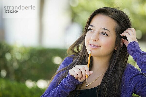 Attraktive nachdenkliche gemischtrassige Studentin mit Bleistift auf Campus-Bank sitzend