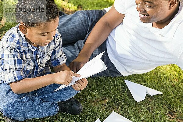 Glücklicher gemischtrassiger Vater und Sohn spielen mit Papierflugzeugen im Park