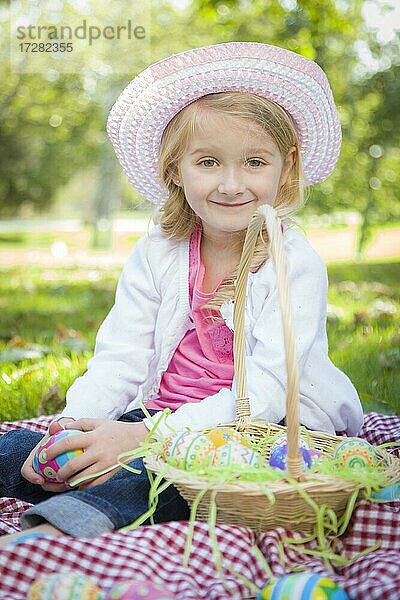 Nettes junges Mädchen auf Picknick-Decke trägt Hut genießt ihre Ostereier draußen im Park