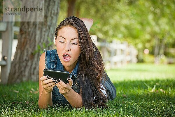 Schockierte gemischtrassige junge Frau textet auf ihrem Handy draußen im Gras liegend