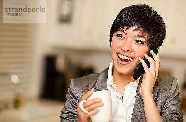 Hübsche lächelnde multiethnische Frau mit Kaffee und sprechen auf einem Handy in ihrer Küche