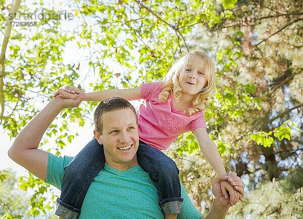 Nettes junges Mädchen reitet Huckepack auf den Schultern ihres Vaters draußen im Park