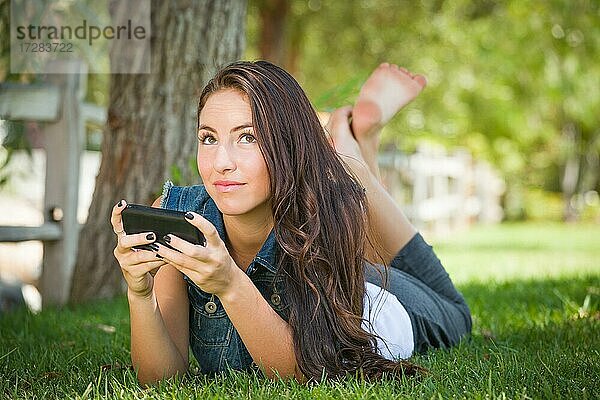 Attraktive glückliche gemischtrassige junge Frau Texting auf ihrem Handy draußen im Gras liegend