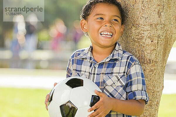 Gemischtrassiger Junge hält Fußball im Park gegen einen Baum