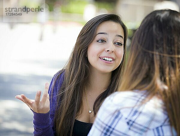 Attraktive ausdrucksstarke junge gemischtrassige Studentin sitzt und spricht mit Freundin draußen auf der Bank
