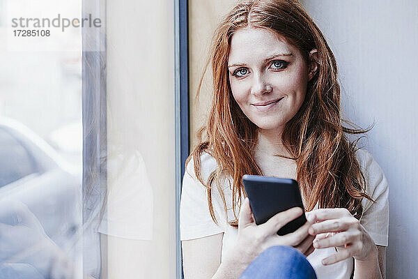 Lächelnde Frau  die ihr Handy hält  während sie zu Hause am Fenster sitzt
