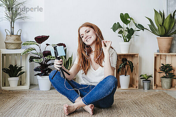 Lächelnde Frau mit Kopfhörern während eines Videoanrufs  während sie zu Hause auf dem Boden sitzt