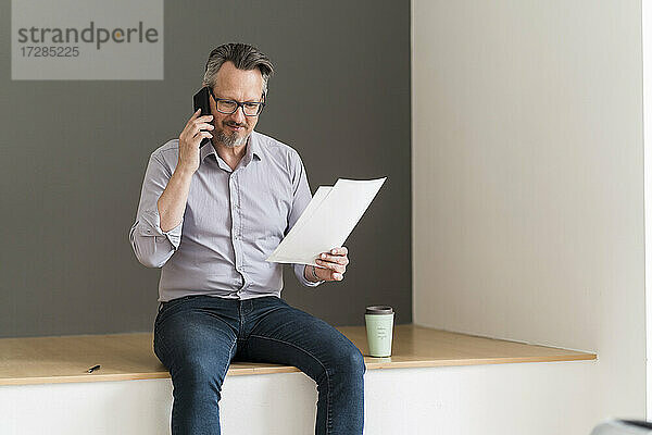 Männlicher Unternehmer mit Dokument  der im Büro mit einem Mobiltelefon spricht