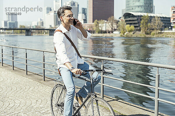 Lächelnder Geschäftsmann auf dem Fahrrad  während er am Fluss in der Stadt mit seinem Handy telefoniert