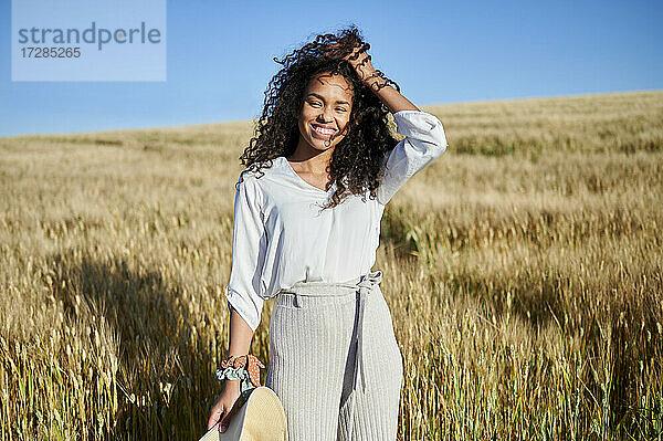 Lächelnde junge Frau steht mit der Hand im Haar in einem Weizenfeld an einem sonnigen Tag