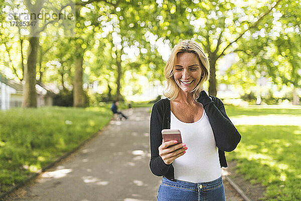 Glückliche reife Frau mit Hand im Haar  die ein Mobiltelefon in einem öffentlichen Park benutzt