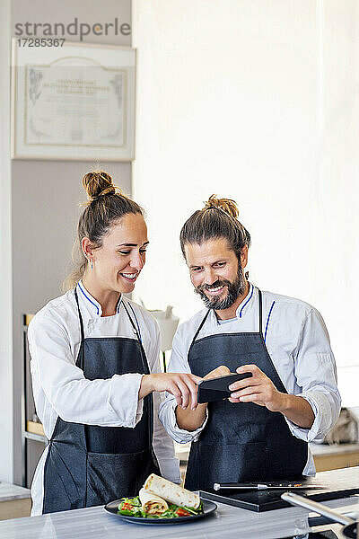 Lächelnde weibliche und männliche Köche  die sich in einem Restaurant über ein Mobiltelefon unterhalten