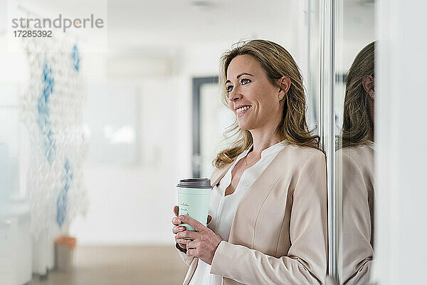 Lächelnde Unternehmerin mit wiederverwendbarem Kaffeebecher  der an einer schalldichten Kabine im Büro lehnt
