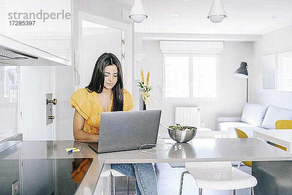 Frau benutzt Laptop  während sie neben einer Schüssel auf dem Küchentisch sitzt