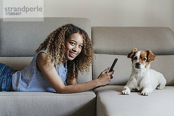 Lockenhaarige Frau mit Smartphone auf Sofa liegend neben Hund im Wohnzimmer