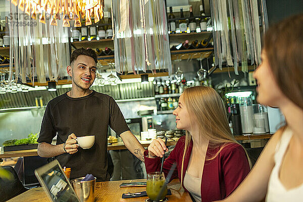 Lächelnder junger Mann mit Kaffeetasse im Gespräch mit Freundinnen am Tisch in einer Bar