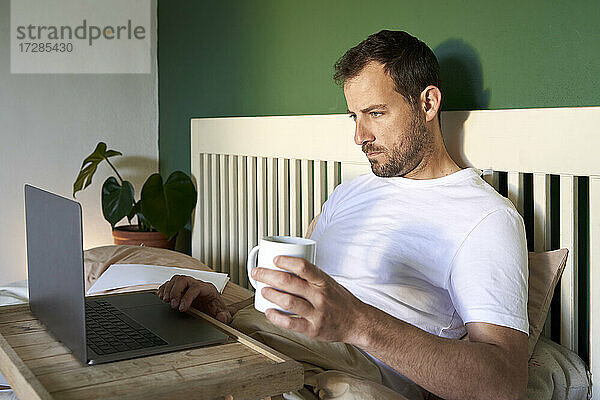 Mann mit Kaffeetasse und Laptop  während er sich zu Hause ausruht