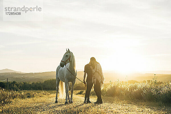 Junges Paar  das sich umarmt  während es bei Sonnenuntergang neben einem Pferd auf einem Feld steht