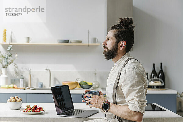 Nachdenklicher Mann mit Bart  der eine Kaffeetasse hält  während er in der Küche am Laptop sitzt