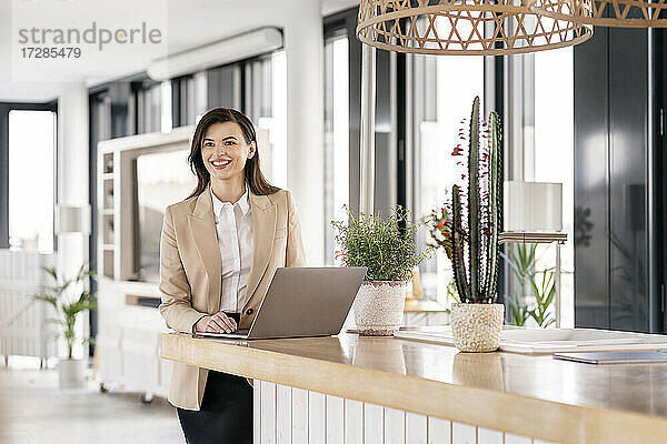 Lächelnde Unternehmerin schaut weg  während sie vor einem Laptop in einer Büro-Cafeteria steht