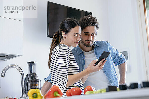 Lächelndes Paar  das in der heimischen Küche gemeinsam ein Mobiltelefon benutzt