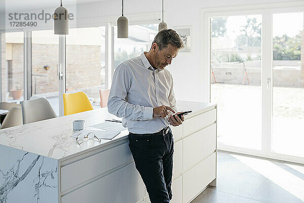 Männlicher Freiberufler arbeitet am Handy  während er zu Hause vor der Kücheninsel steht