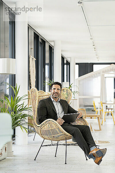 Geschäftsmann mit digitalem Tablet  der wegschaut  während er auf einem Stuhl in einer Büro-Cafeteria sitzt