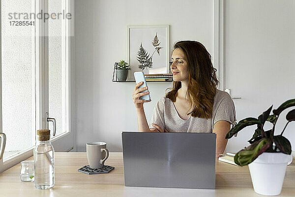 Lächelnde Frau  die ein Smartphone am Schreibtisch im Home Office benutzt