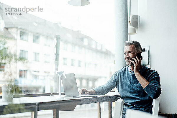 Lächelnder Geschäftsmann mit Laptop  der in einem Café mit seinem Mobiltelefon spricht
