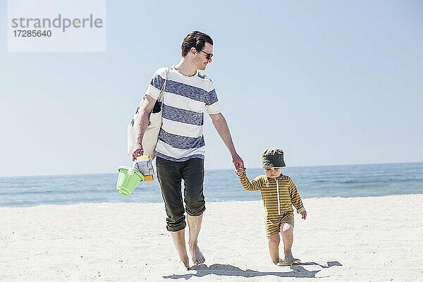 Mann und Sohn halten sich an den Händen und gehen an einem sonnigen Tag am Strand spazieren