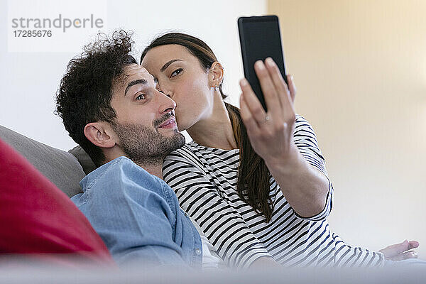 Freundin nimmt Selfie durch Smartphone beim Küssen auf Freund Wange zu Hause