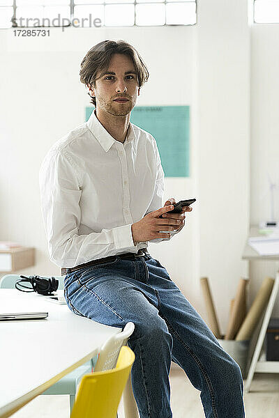 Geschäftsmann  der ein Mobiltelefon hält  während er in einem kreativen Büro auf dem Schreibtisch sitzt