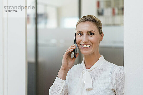 Lächelnde blonde Geschäftsfrau  die mit einem Handy telefoniert und an der Tür eines Büros steht