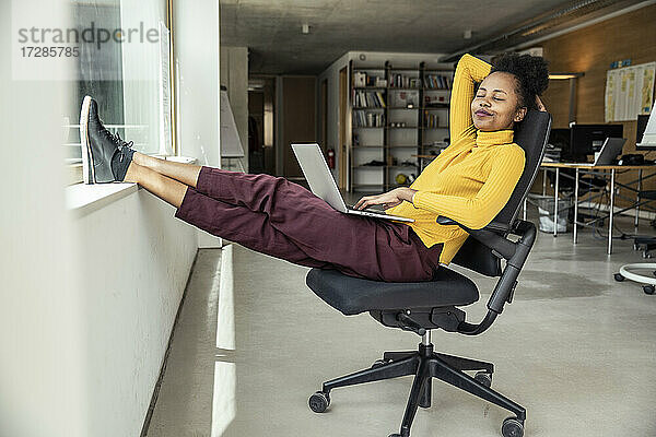 Geschäftsfrau mit Laptop  die sich auf einem Stuhl in einem Büroappartement entspannt